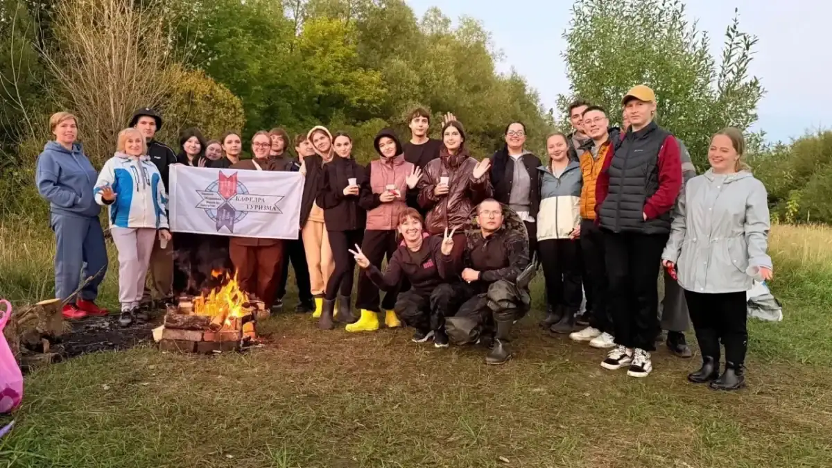 Участники мероприятия стоят у костра с флагом на лесной поляне.