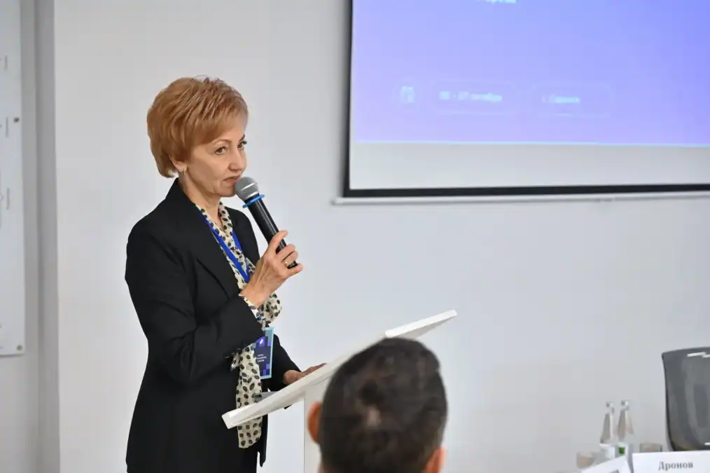 Женщина выступает с микрофоном у экрана с презентацией.