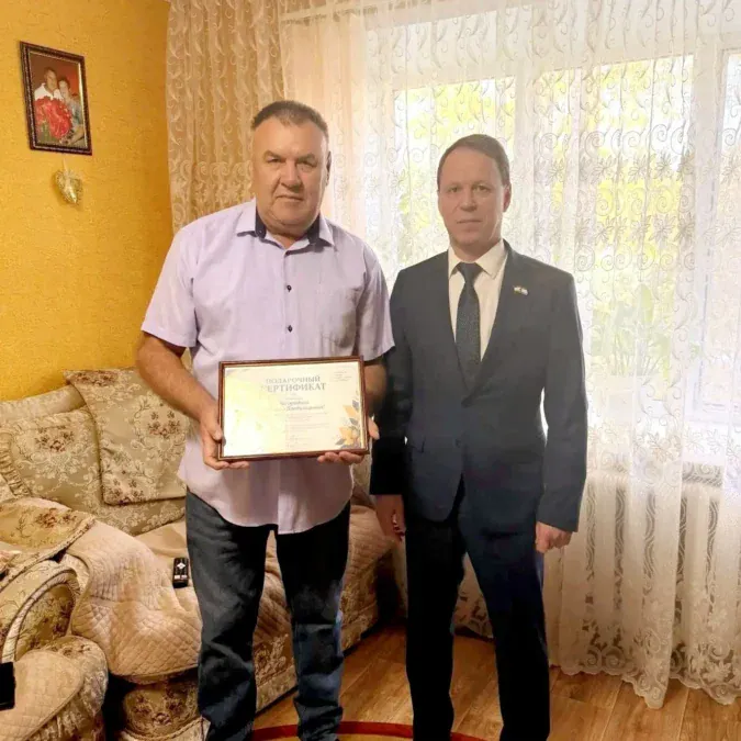 Двое мужчин стоят в комнате, один держит подарочный сертификат.