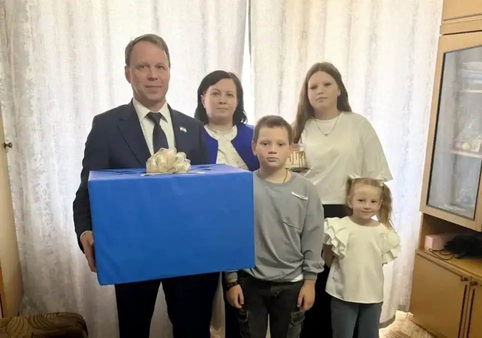 Мужчина с большой синей подарочной коробкой стоит рядом с семьёй и детьми.