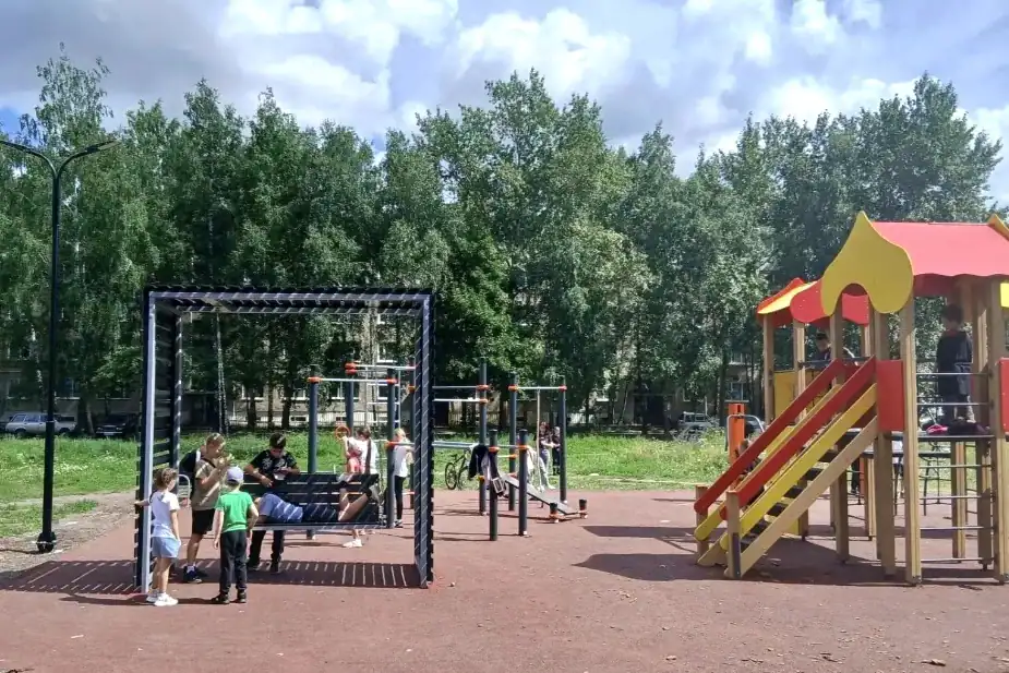 Дети играют у качелей и комплекса с горкой на благоустроенной площадке.