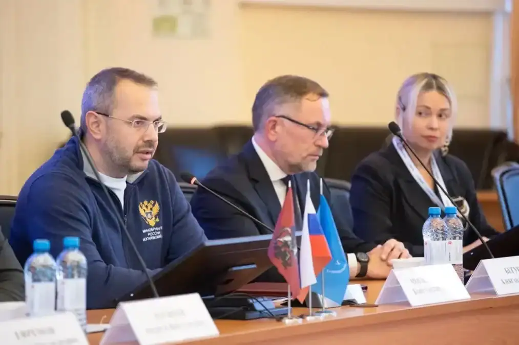 Трое участников заседания сидят за столом с микрофонами и настольными флагами.