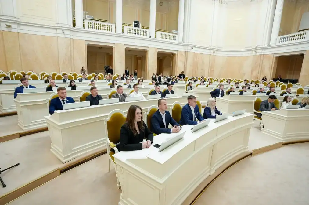 Делегаты сидят за рядами белых столов в парламентском зале.
