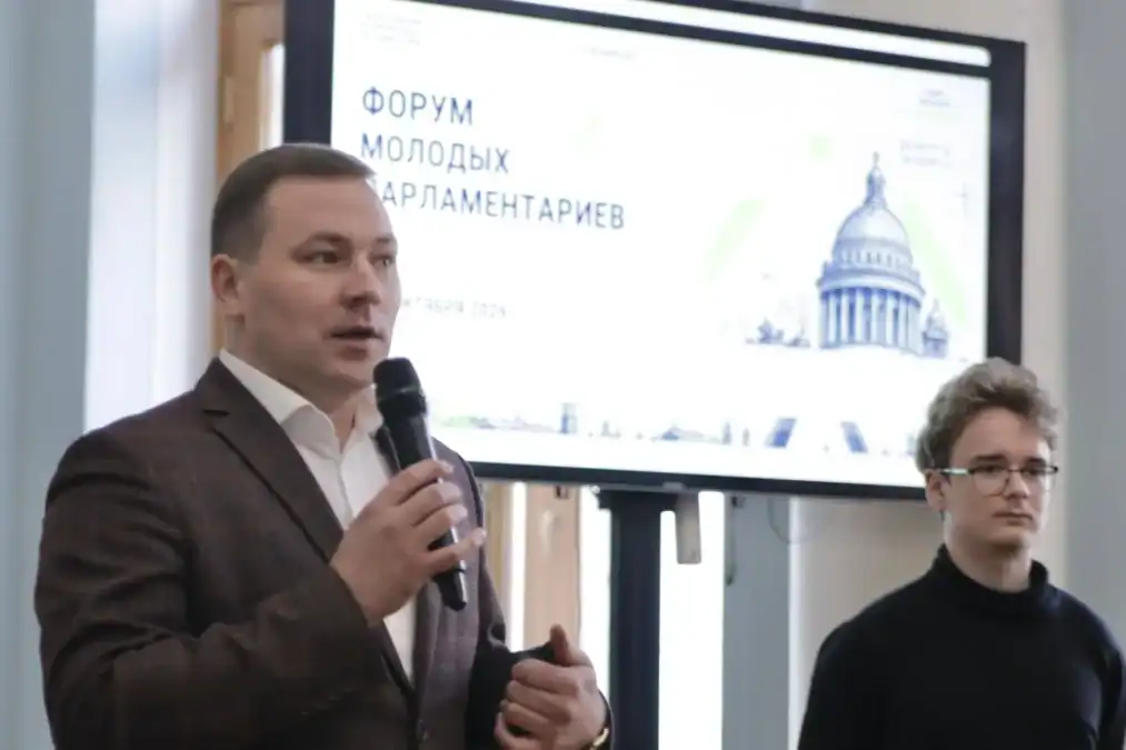 Мужчина выступает с микрофоном перед экраном форума молодых парламентариев.