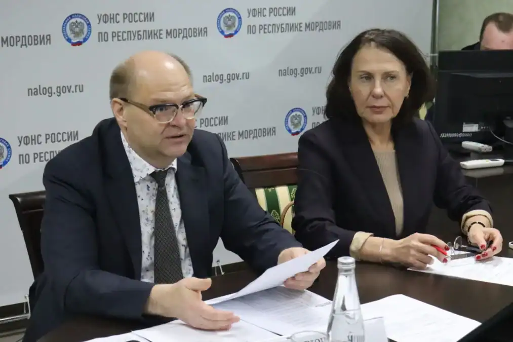 Двое участников заседания сидят за столом с документами и водой.