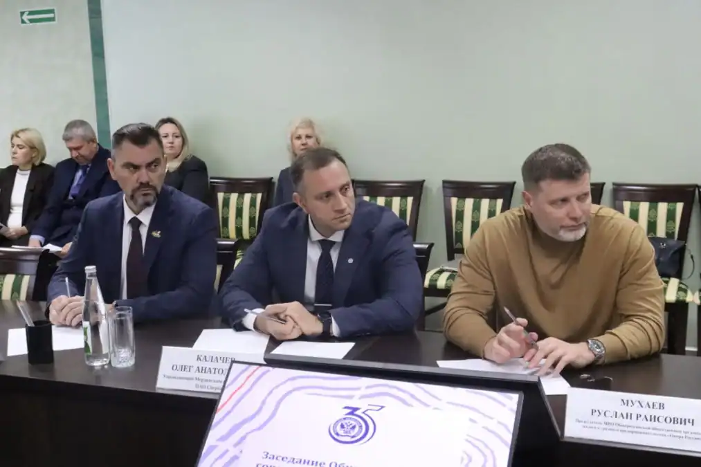 Трое мужчин участвуют в заседании за столом с именными табличками.