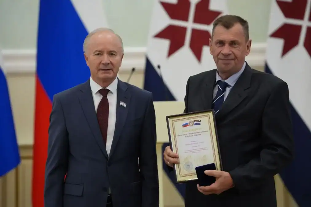 Двое мужчин в костюмах позируют с грамотой на фоне флагов Мордовии.