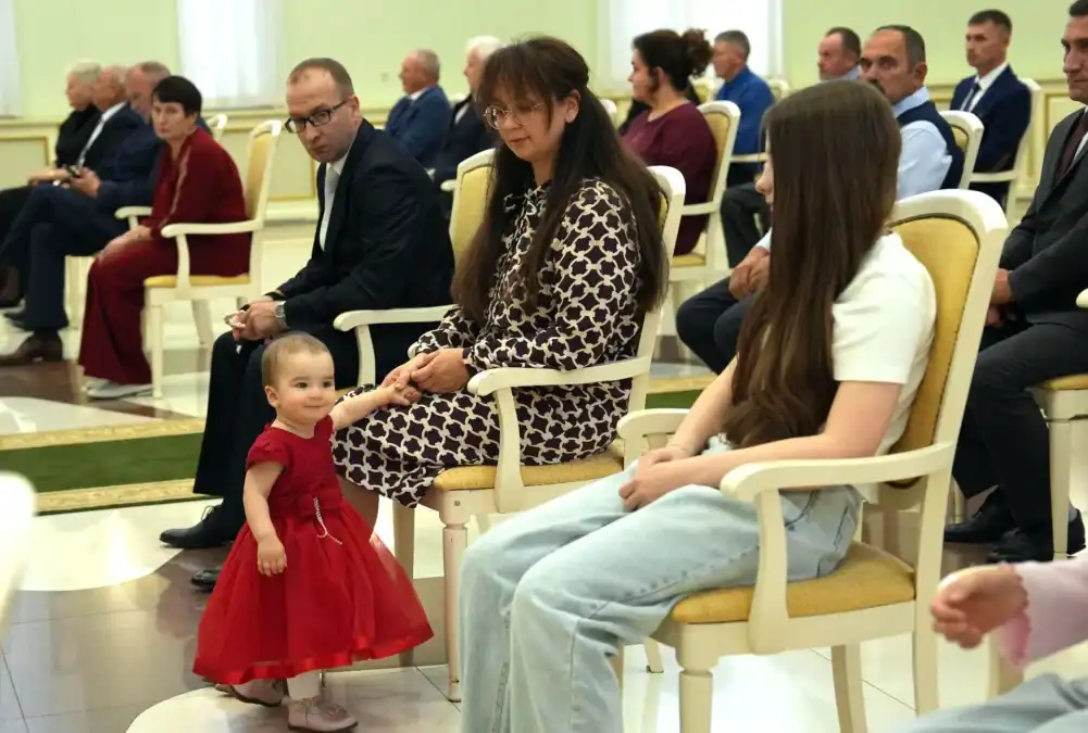 Участники торжественного мероприятия сидят в зале, рядом стоит маленькая девочка в красном платье.