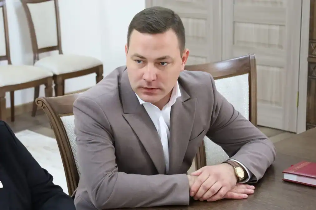 Мужчина в сером костюме сидит за столом рядом с папкой.