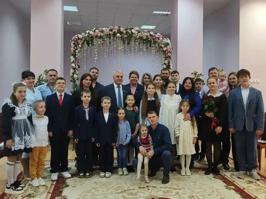 Многодетные семьи с детьми позируют у украшенной цветочной арки в помещении.