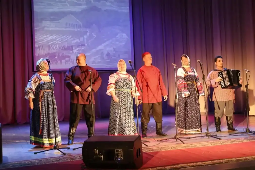 Исполнители в народных костюмах поют на сцене, рядом играет баянист.