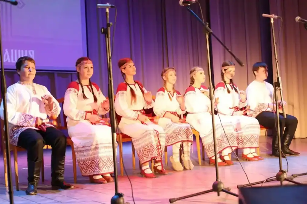 Участники в вышитых народных костюмах сидят на сцене перед микрофонами.