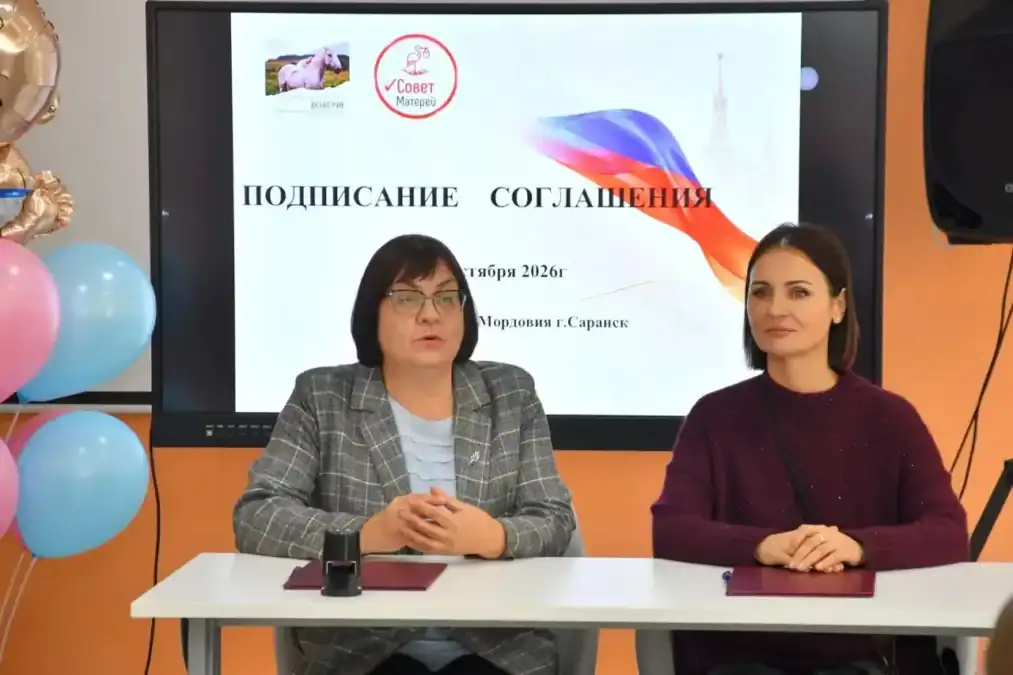 Две женщины сидят за столом перед экраном с надписью о подписании соглашения.