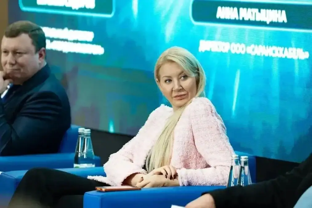 Женщина сидит в синем кресле на фоне экрана мероприятия.