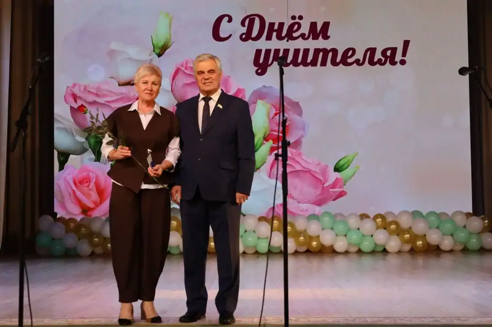 Женщина и мужчина стоят на сцене перед экраном с поздравлением ко Дню учителя.