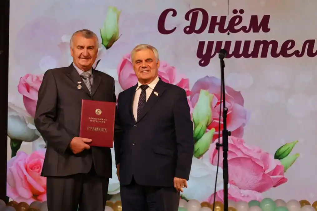 Двое мужчин позируют на сцене, один держит красную папку с гербом.