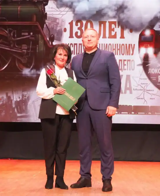 Женщина с букетом и папкой стоит рядом с мужчиной на сцене.
