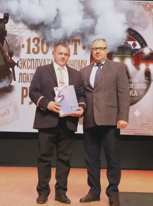 Двое мужчин стоят на сцене, один держит синюю папку.