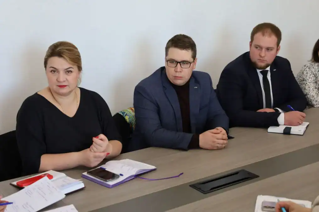 Трое участников совещания сидят за столом с документами и записями.