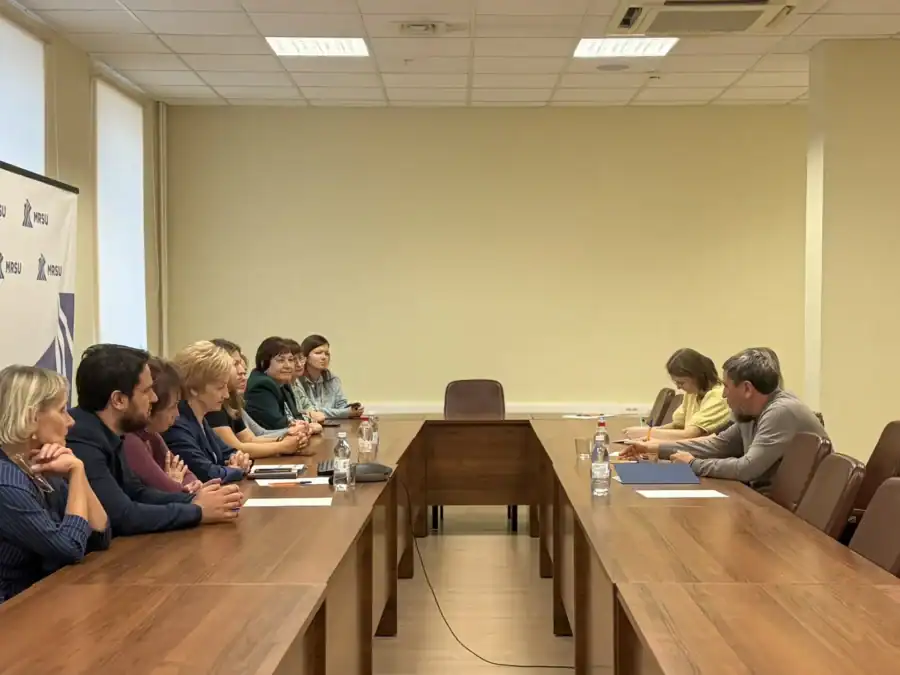 Участники встречи сидят за длинным столом с документами и водой.