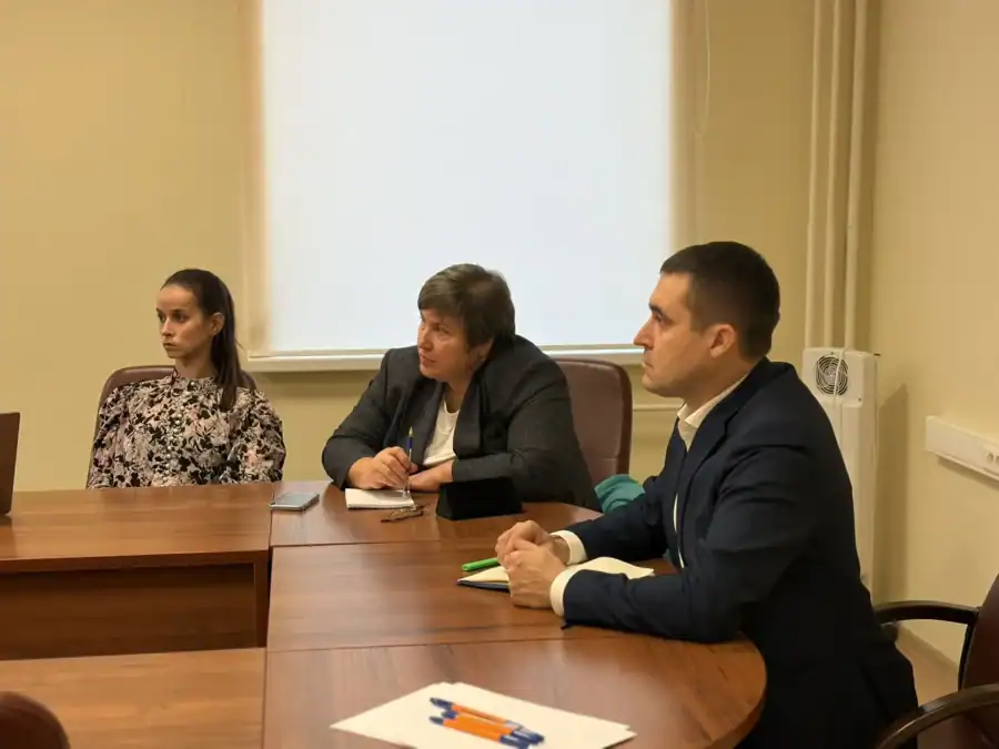 Трое участников совещания сидят за столом с блокнотами и документами.