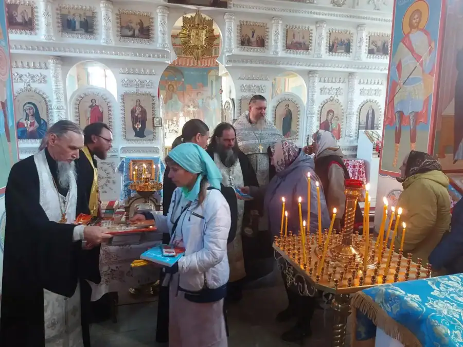 Священнослужитель за церковным столом общается с женщинами, рядом горят свечи.
