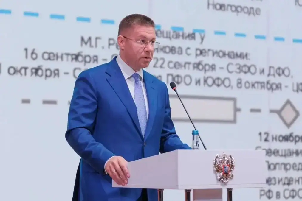 Мужчина в синем костюме выступает у трибуны перед экраном со схемой.