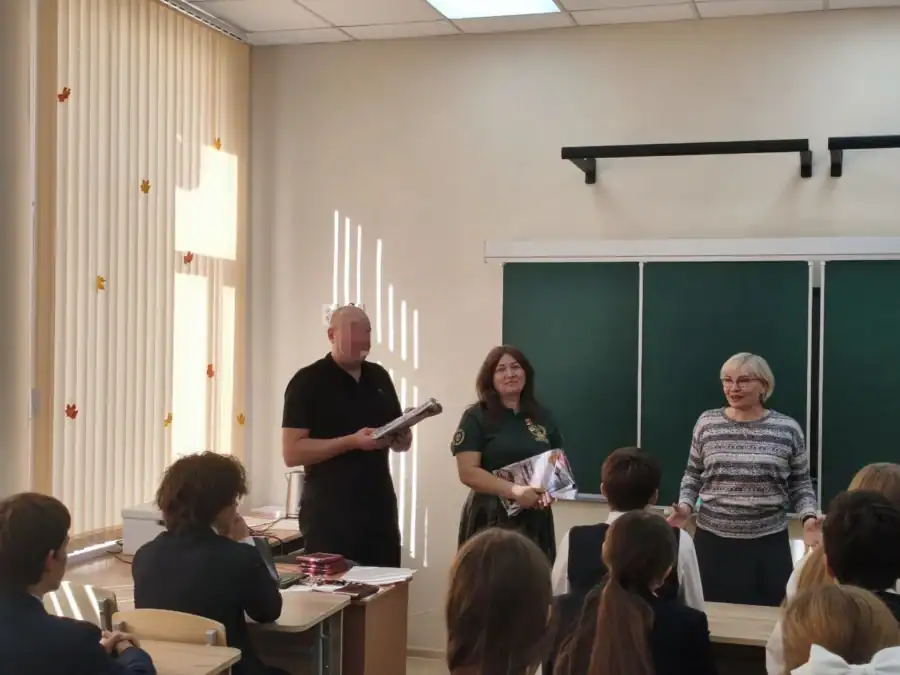 Трое взрослых стоят перед сидящими школьниками у доски в классе.