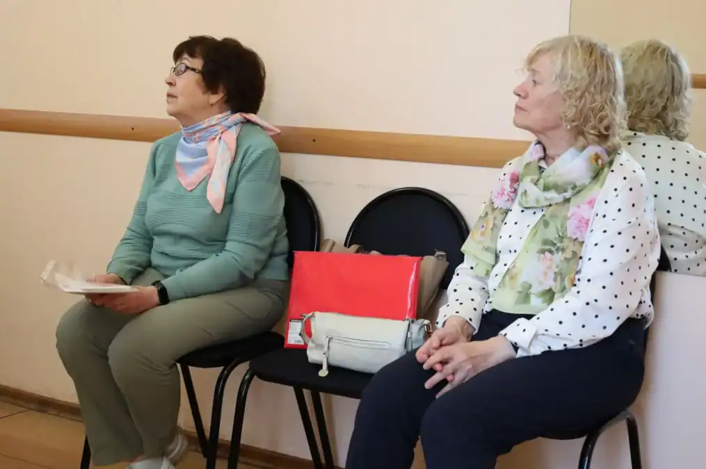 Две женщины ожидают в коридоре на стульях, одна держит бумаги.