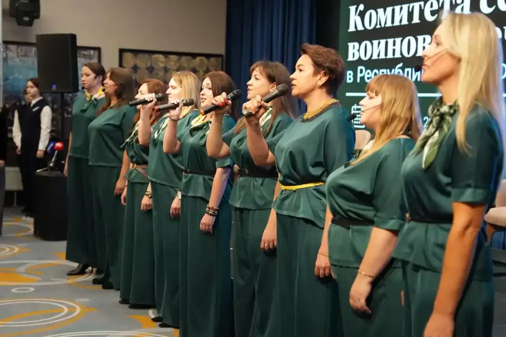 Женщины в зелёных костюмах поют в микрофоны перед экраном форума.