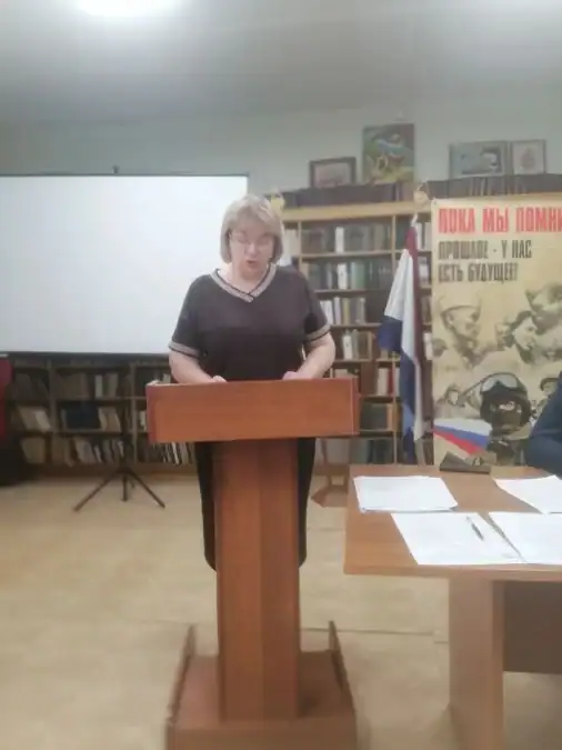 Женщина читает документы за трибуной в зале с книжными полками.
