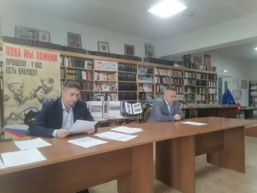Двое мужчин сидят за длинным столом с разложенными документами в библиотечном зале.