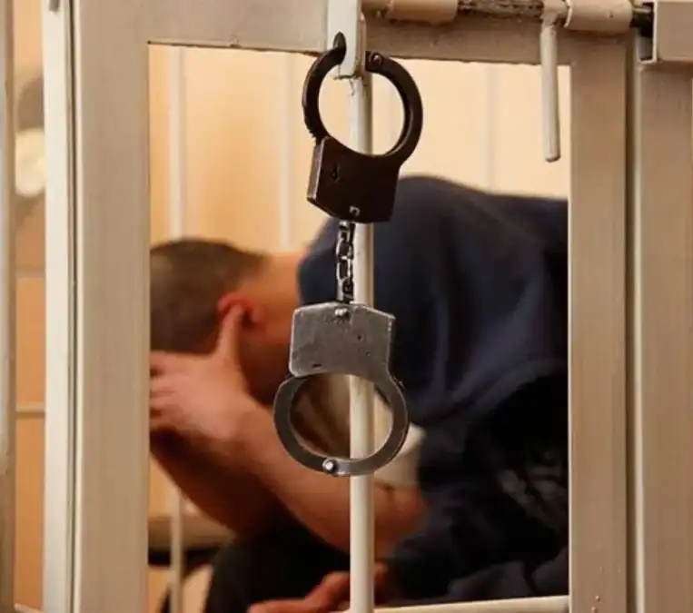 Наручники висят на металлической решётке перед сидящим мужчиной.