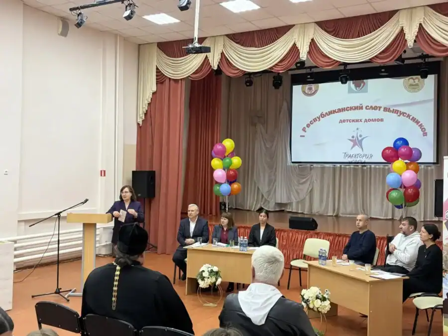 Участники встречи сидят за столами перед сценой с экраном и шарами.
