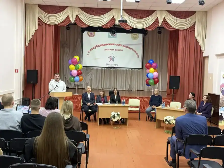 Спикер выступает перед участниками слёта в зале с праздничным оформлением сцены.