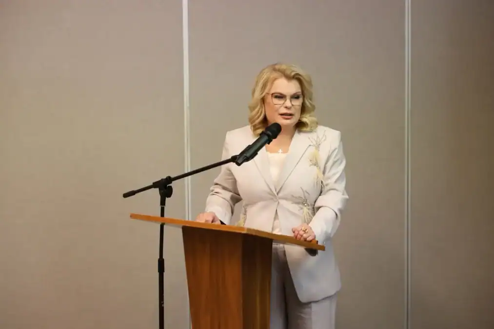 Женщина в светлом костюме выступает с трибуны у микрофона.