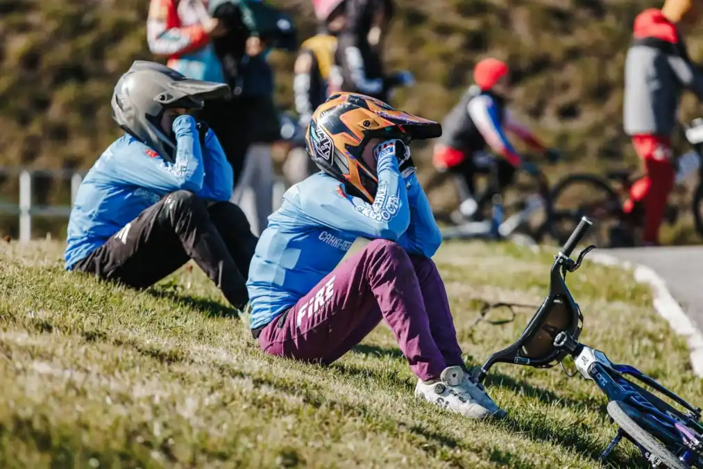 Два участника в шлемах сидят на траве рядом с BMX-велосипедом.