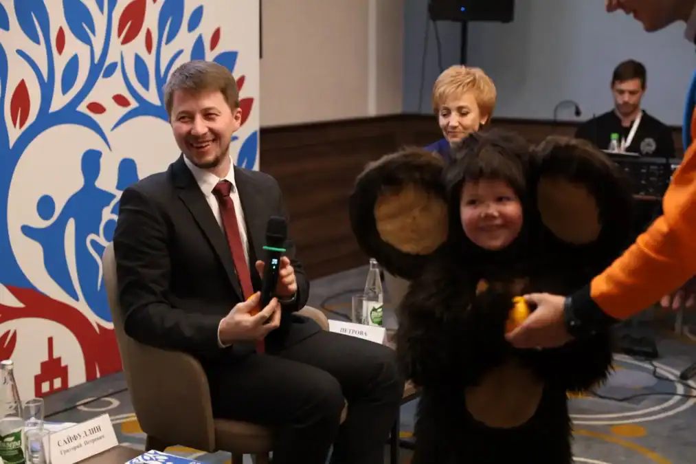 Мужчина с микрофоном сидит рядом с ребёнком в костюме Чебурашки.