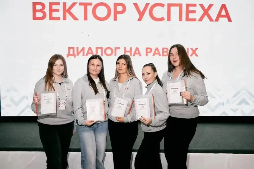 Пять участниц в серых толстовках держат сертификаты перед экраном проекта.