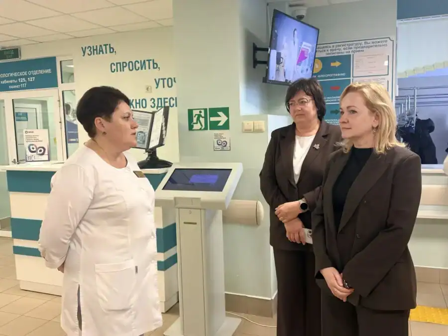 Сотрудница поликлиники беседует с двумя посетительницами у стойки регистрации.