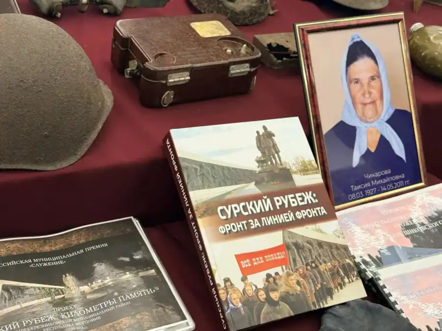 На столе лежат книги, каска, чемодан и портрет пожилой женщины.