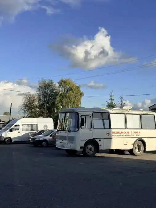 Белый медицинский автобус стоит на площадке рядом с легковыми автомобилями.