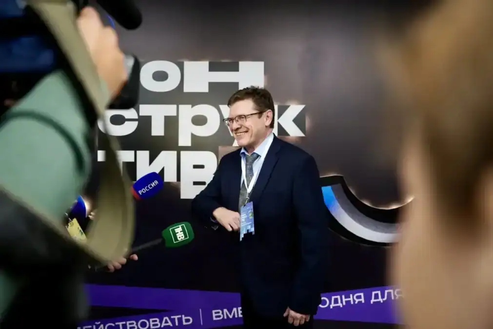 Мужчина с бейджем отвечает журналистам перед брендированной стеной форума.