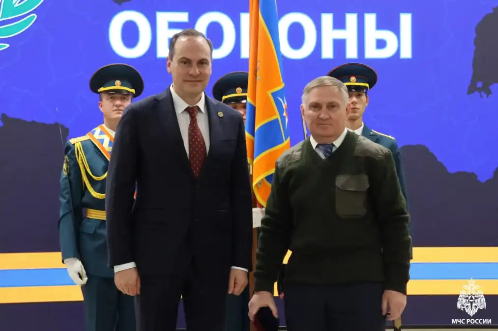 Двое мужчин стоят перед почётным караулом и флагом на сцене.