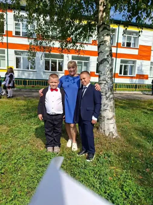 Бабушка в синем платье стоит с двумя мальчиками у здания.