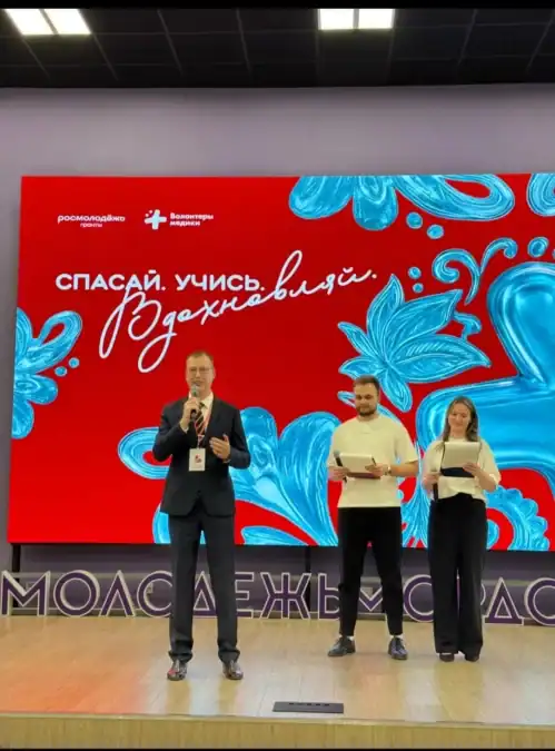 Мужчина в костюме говорит в микрофон, рядом стоят два участника мероприятия.