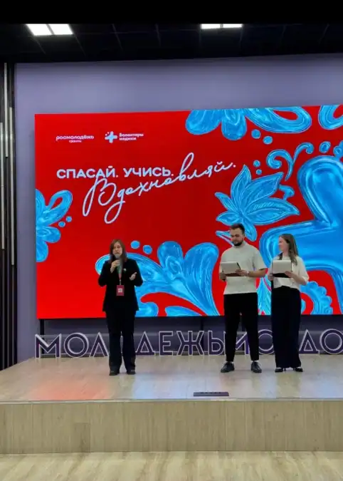 Женщина выступает с микрофоном, двое участников держат листы на сцене.