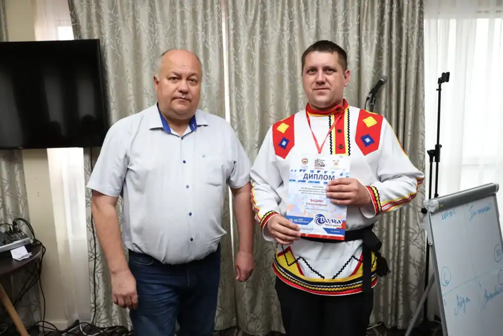 Мужчина в традиционном костюме держит диплом рядом с другим участником.