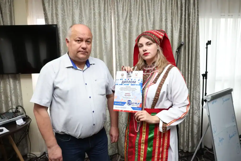 Участница в народном костюме держит диплом рядом с мужчиной.