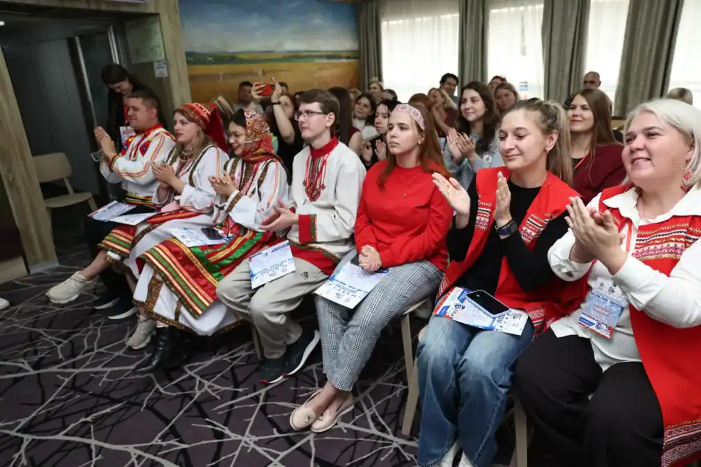 Участники в традиционных костюмах и повседневной одежде аплодируют в зале.